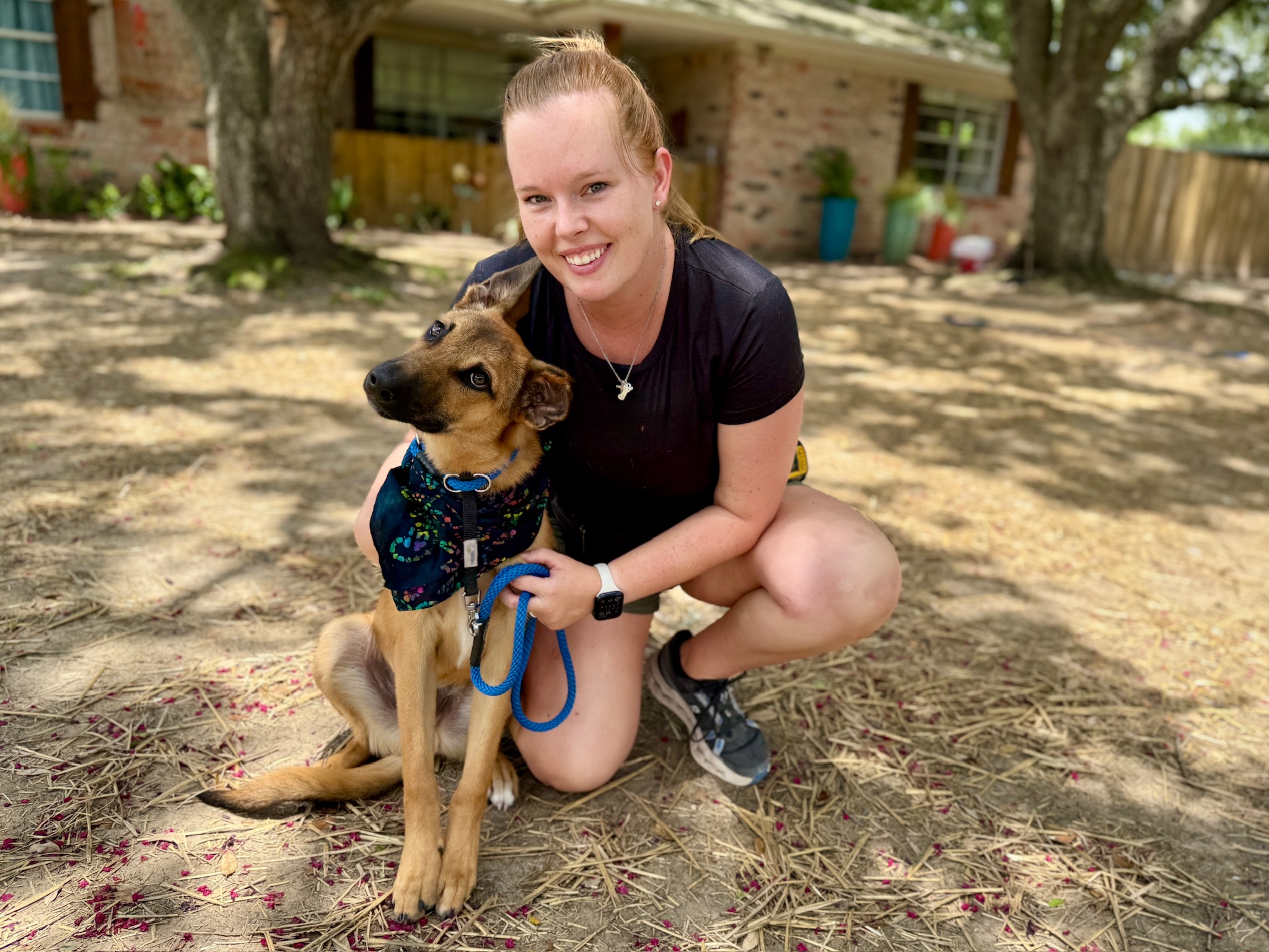 A woman kneeling beside a tan dog wearing a harness in an outdoor yard