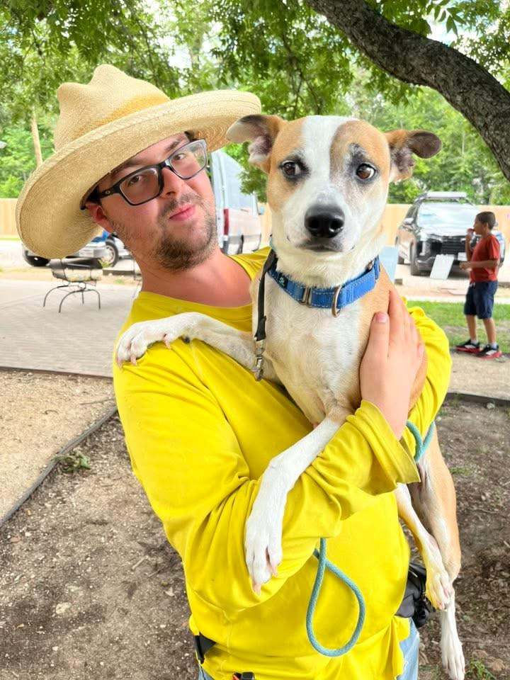 A man wearing a cowboy hat holding a tan-and-white dog outdoors
