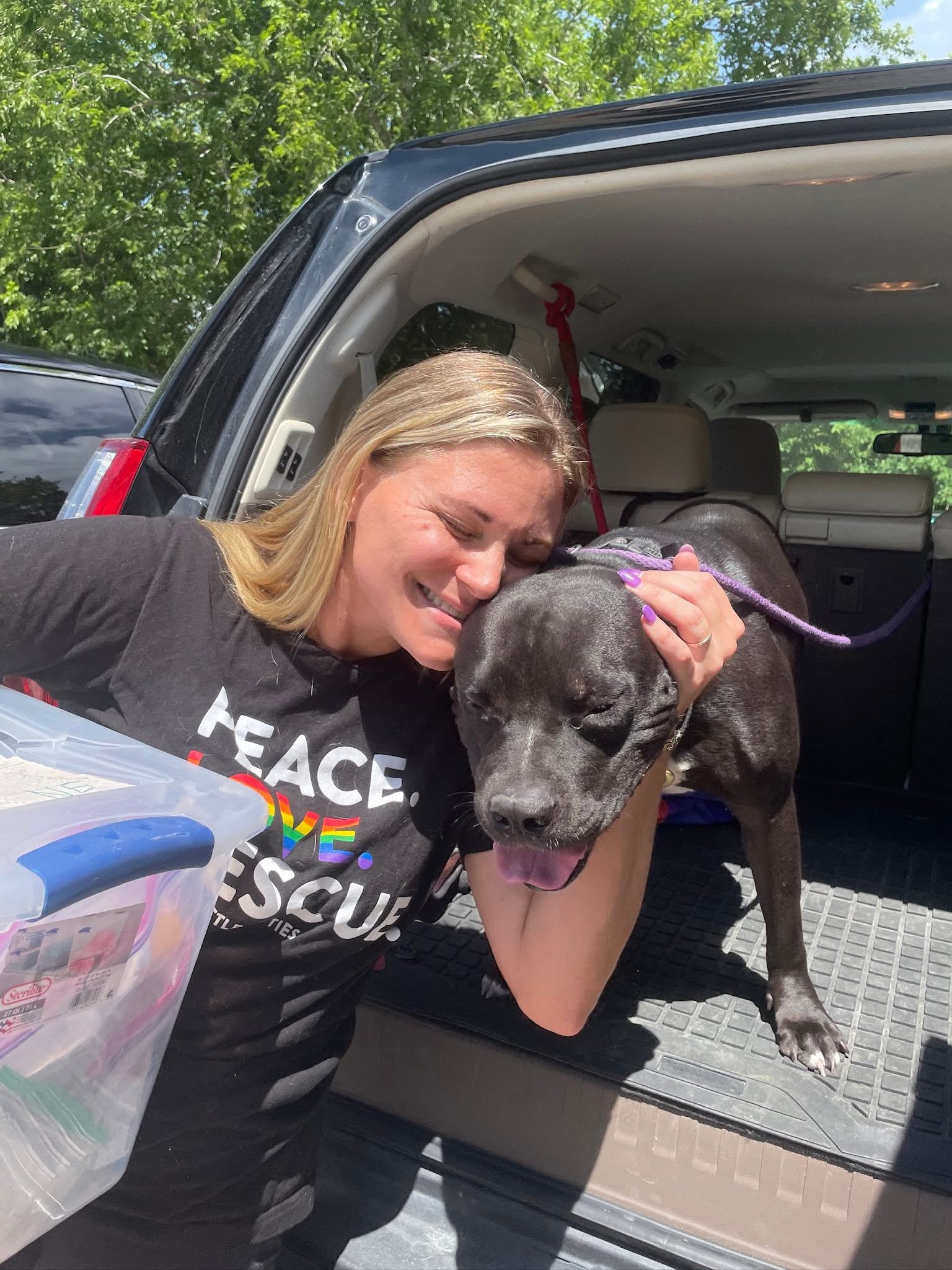 A foster caregiver kissing a gray dog beside an open vehicle